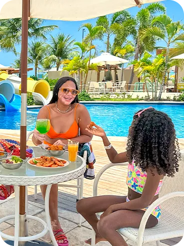 All inclusive à beira da piscina Três pessoas sentadas à mesa redonda sob ombrelone, com petiscos e bebidas; piscina e toboáguas azuis ao fundo, palmeiras e céu claro. Um homem segura uma cerveja, a mulher um drink verde e a criança prova um salgado.