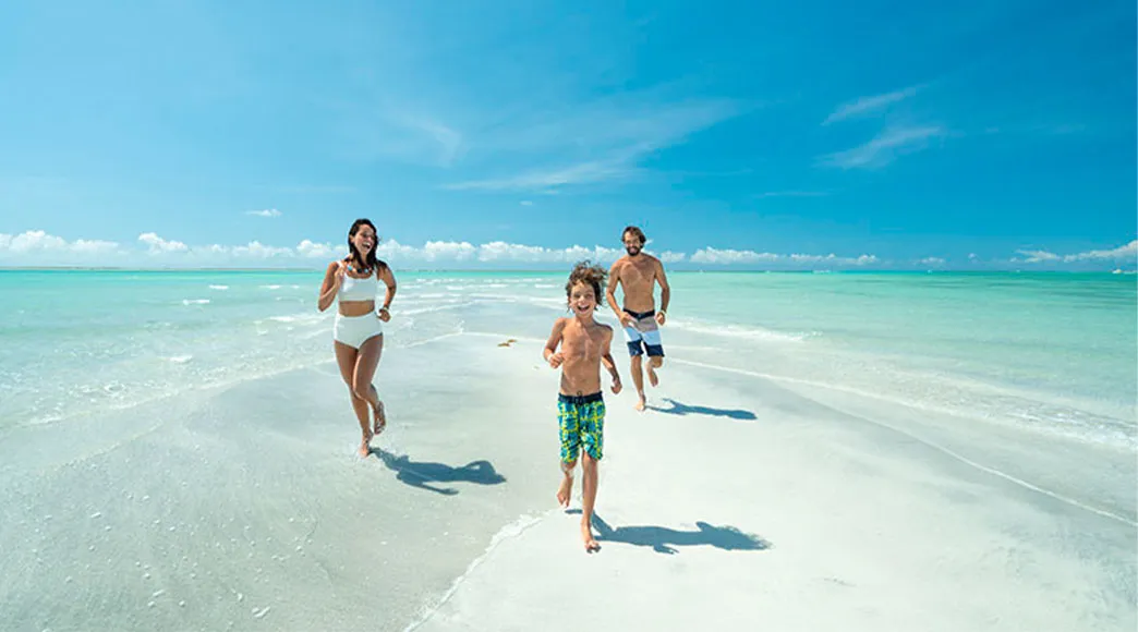 Família em passeio de lancha em Maragogi Uma família com pai, mãe e filho sentados na proa de uma lancha, sorrindo, durante um passeio nas águas cristalinas de cor verde-esmeralda das Galés de Maragogi, com a orla ao fundo.