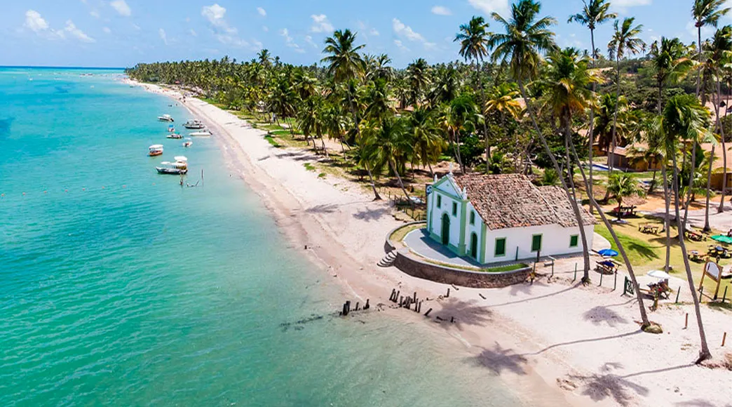 Capela de São Benedito na Praia dos Carneiros Vista aérea da Capela de São Benedito, uma igrejinha branca localizada na areia da Praia dos Carneiros, cercada por um extenso coqueiral e pelo mar calmo de águas esverdeadas com barcos ancorados.