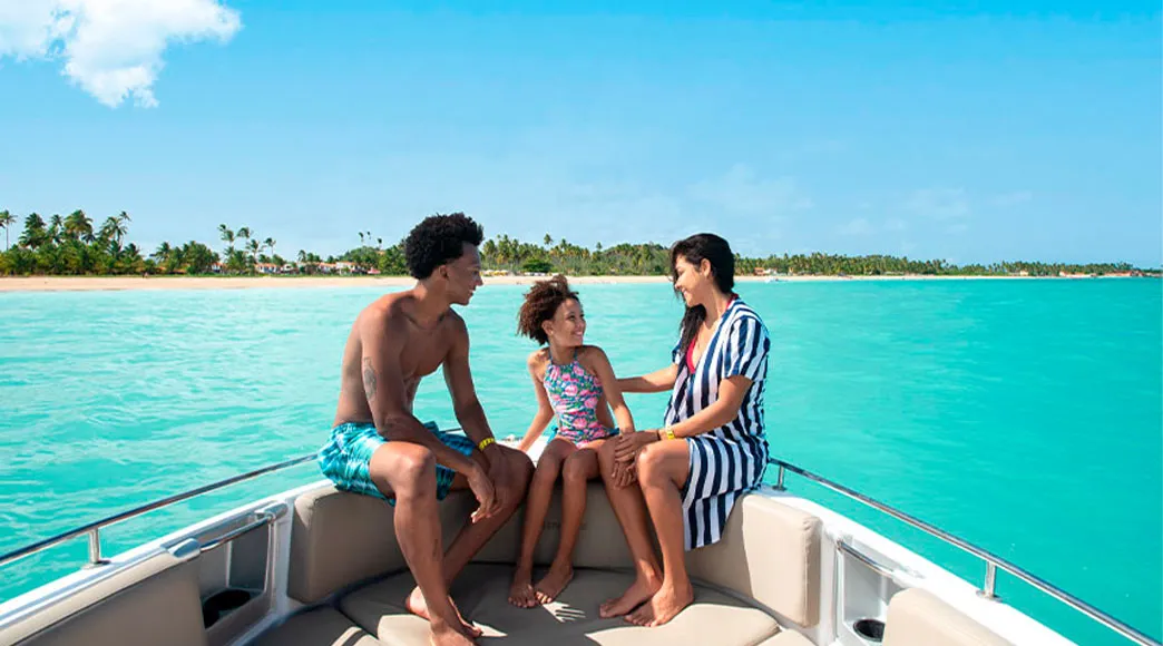 Passeio de barco na Praia de Burgalhau Família com pai, mãe e filha sentados na proa de uma lancha, conversando e sorrindo durante um passeio no mar de águas calmas e azul-turquesa, com a orla da Praia de Burgalhau e seus coqueiros ao fundo.