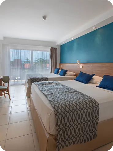 apartamento superior Duas camas queen size dispostas lado a lado, com duas luminárias, sendo uma para cada cama, em frente a painel de madeira com TV a cabo, mesa e cadeira.