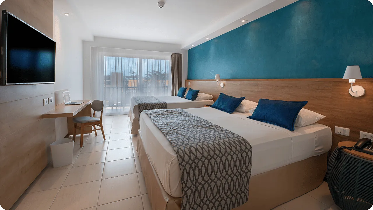 apartamento superior Duas camas queen size dispostas lado a lado, com duas luminárias, sendo uma para cada cama, em frente a painel de madeira com TV a cabo, mesa e cadeira.