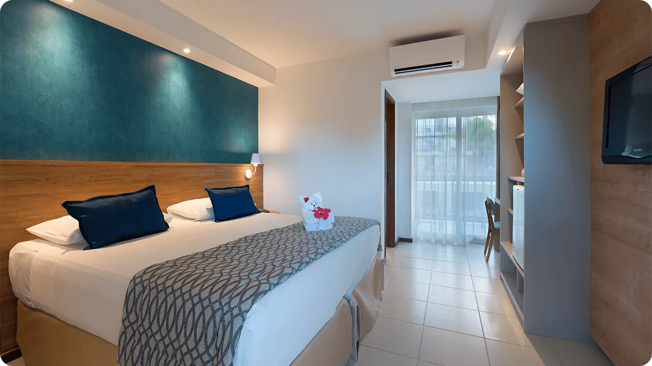 acomodação standard Cama queen size na acomodação Standard, decorada com ursinho  feito de toalha, em frente a roupeiro e painel de madeira com TV a cabo, mesa e cadeira.