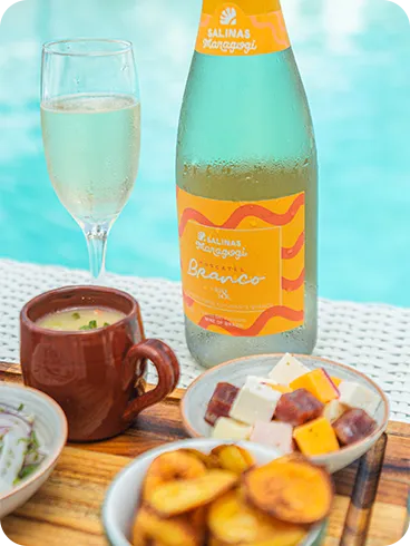 All inclusive com espumante Garrafa de espumante do Salinas Maragogi e taça cheia sobre mesa à beira da piscina, com petiscos variados em tigelas sobre uma tábua.