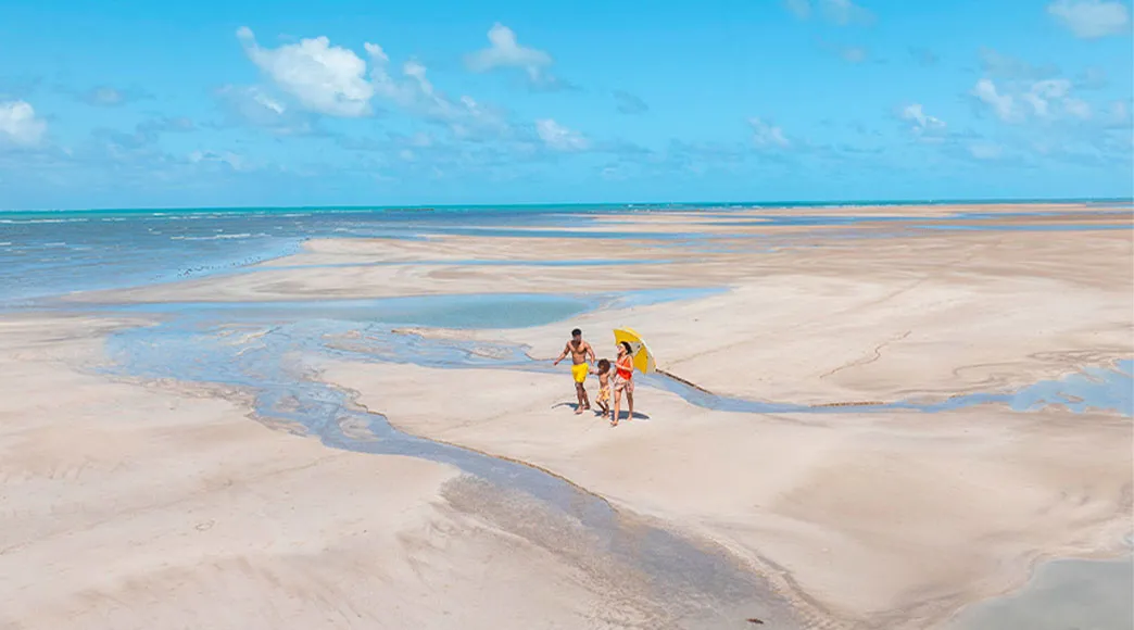 Praia de Maragogi com passeio familiar Família caminhando pela praia de Maragogi, com céu azul, águas cristalinas e um guarda-sol
