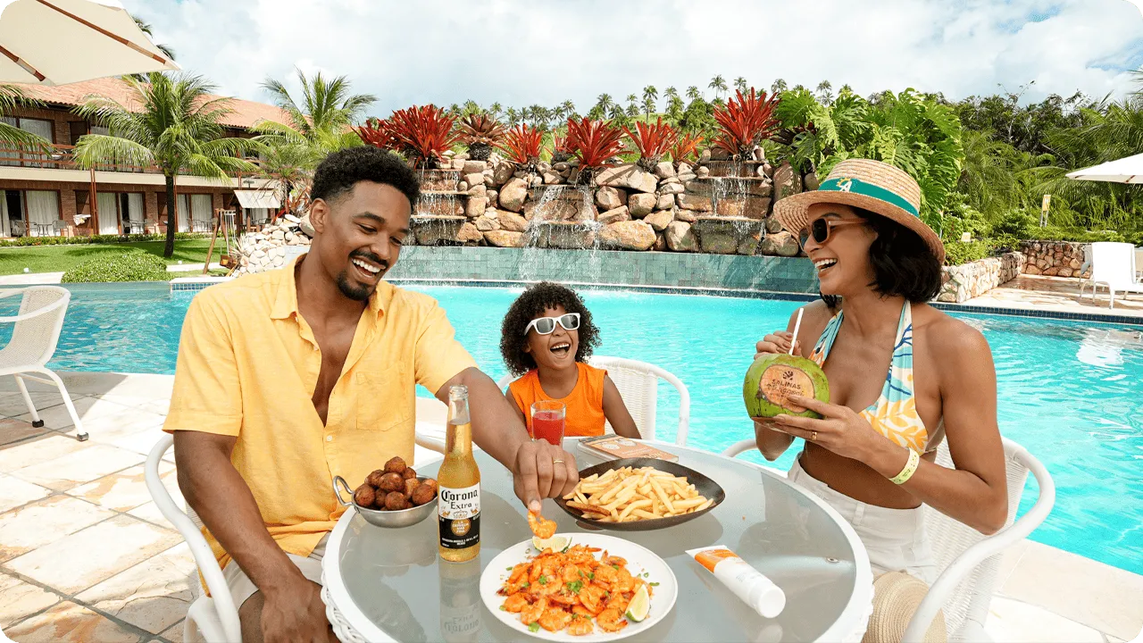 Bocadillos familiares junto a la piscina en Salinas Maragogi Una familia feliz con padre, madre e hija disfrutando de bocadillos y bebidas en una mesa junto a la piscina en Salinas Maragogi, con camarones, papas fritas y coco.