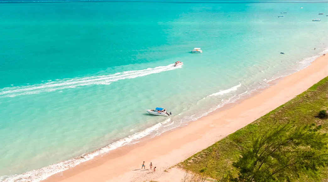 Vista aérea da Praia de Burgalhau Vista aérea da Praia de Burgalhau, com sua longa faixa de areia e vegetação costeira. O mar exibe tons de azul-turquesa, com uma lancha navegando em velocidade e deixando um rastro branco na água.