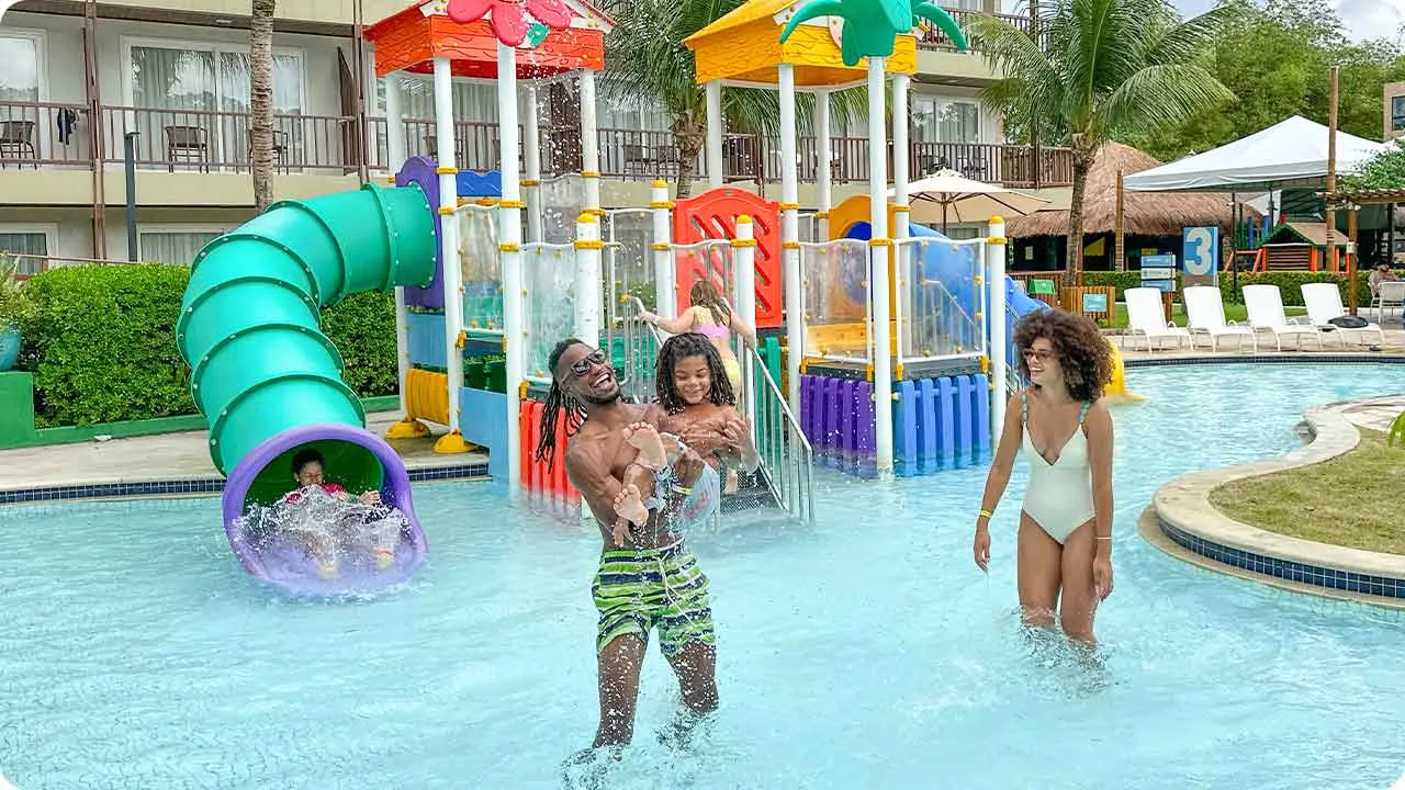Resort pra família Família brincando no parque aquático infantil do Salinas Maceió, com toboágua verde, jatos d’água e piscina rasa.