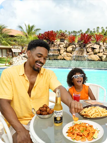 Bocadillos familiares junto a la piscina en Salinas Maragogi Una familia feliz con padre, madre e hija disfrutando de bocadillos y bebidas en una mesa junto a la piscina en Salinas Maragogi, con camarones, papas fritas y coco.