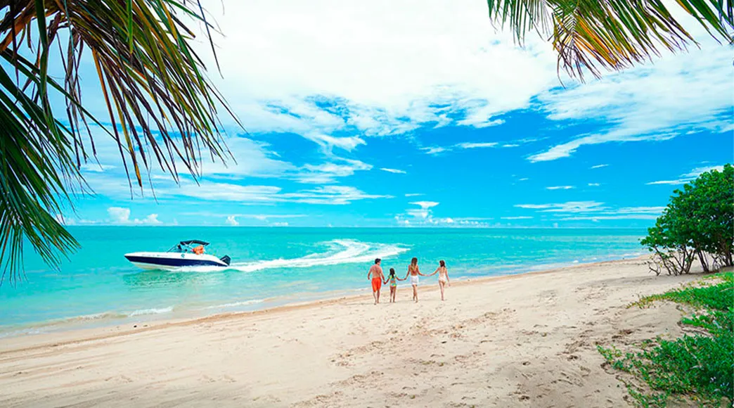 Família caminhando na Praia de Antunes Uma família com pai, mãe e dois filhos caminha de mãos dadas na faixa de areia da Praia de Antunes, com o mar calmo e azul-turquesa à esquerda e uma vegetação verde à direita, enquanto uma lancha navega ao fundo.