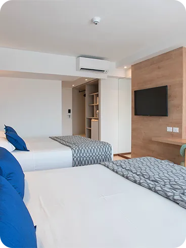 acomodação superior Porta de entrada da acomodação superior ao lado do roupeiro e com duas camas queen size dispostas lado a lado, duas luminárias, sendo uma para cada cama, em frente a painel de madeira com TV a cabo, mesa e cadeira.