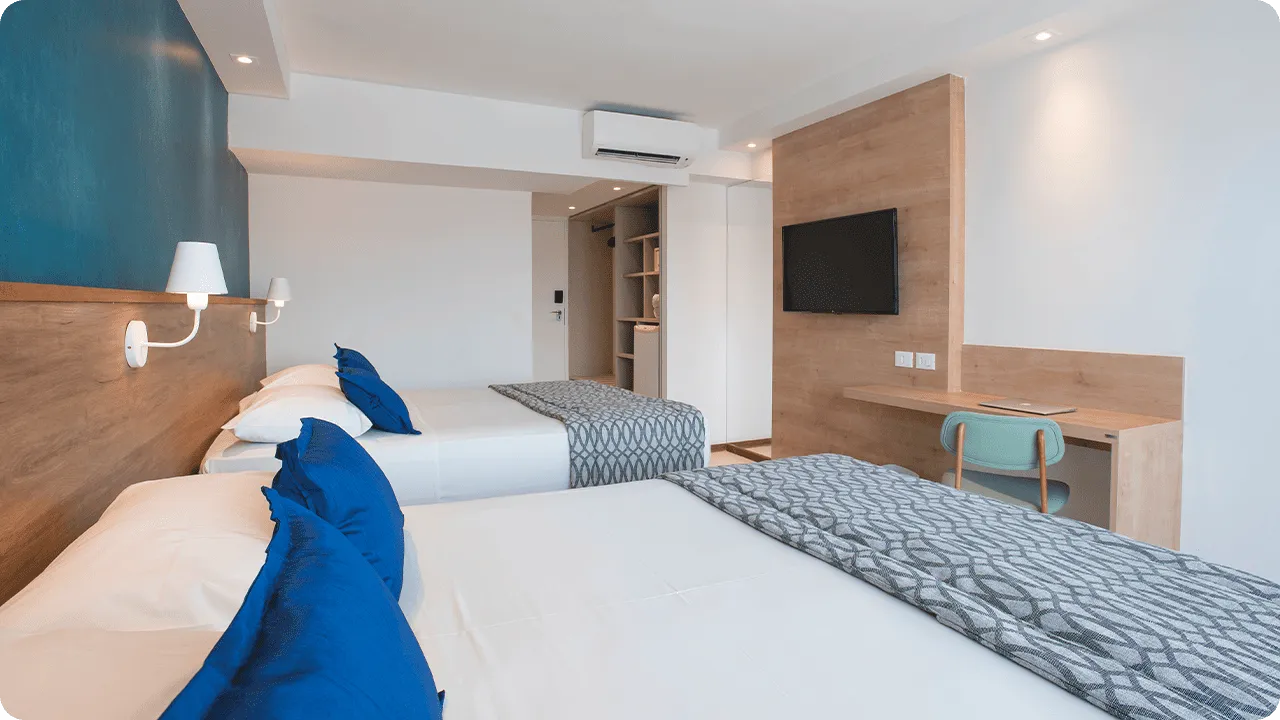 acomodação superior Porta de entrada da acomodação superior ao lado do roupeiro e com duas camas queen size dispostas lado a lado, duas luminárias, sendo uma para cada cama, em frente a painel de madeira com TV a cabo, mesa e cadeira.
