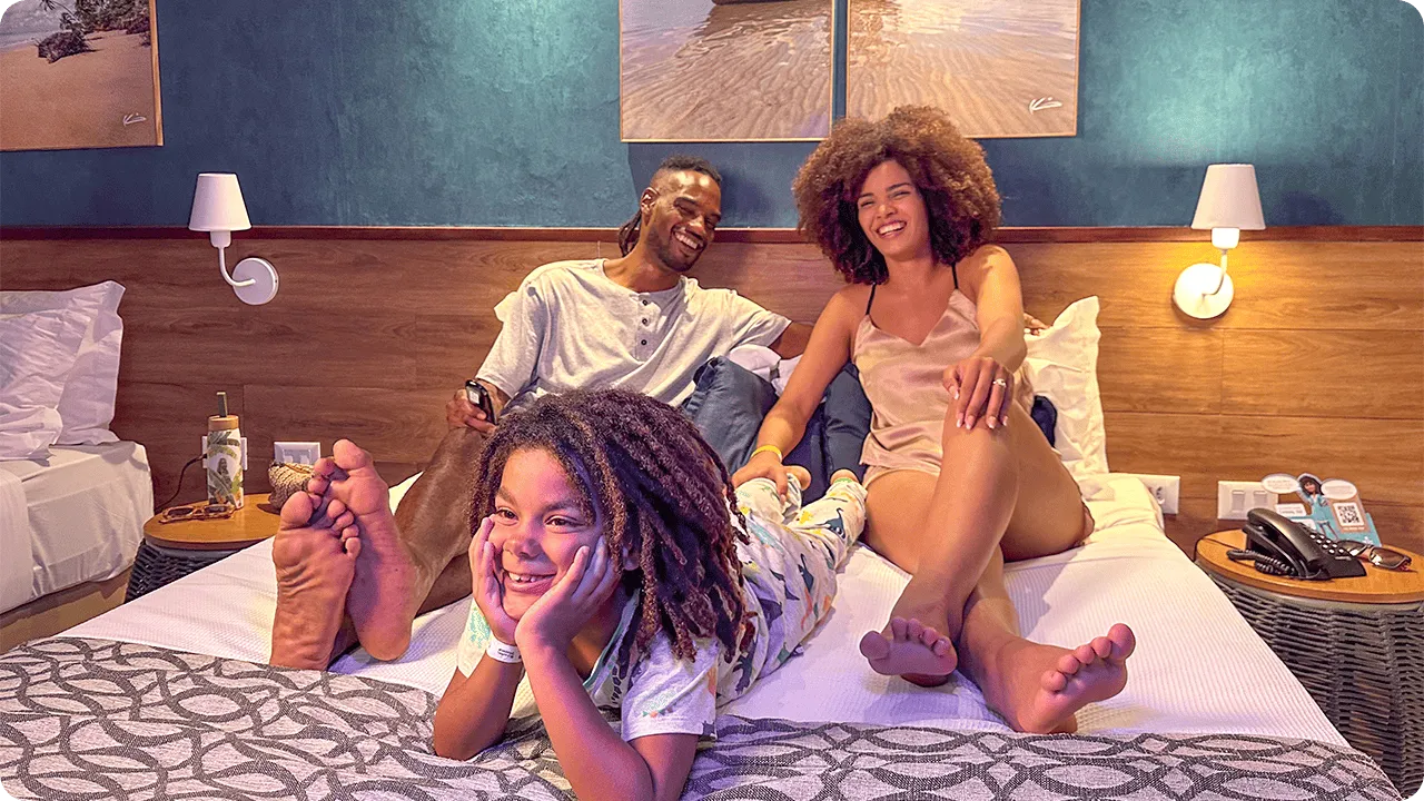 Família em Quarto Standard Família feliz relaxando na cama do quarto Standard, com o filho deitado em primeiro plano e os pais sentados atrás. A decoração do quarto inclui uma parede de destaque azul e quadros de praia.