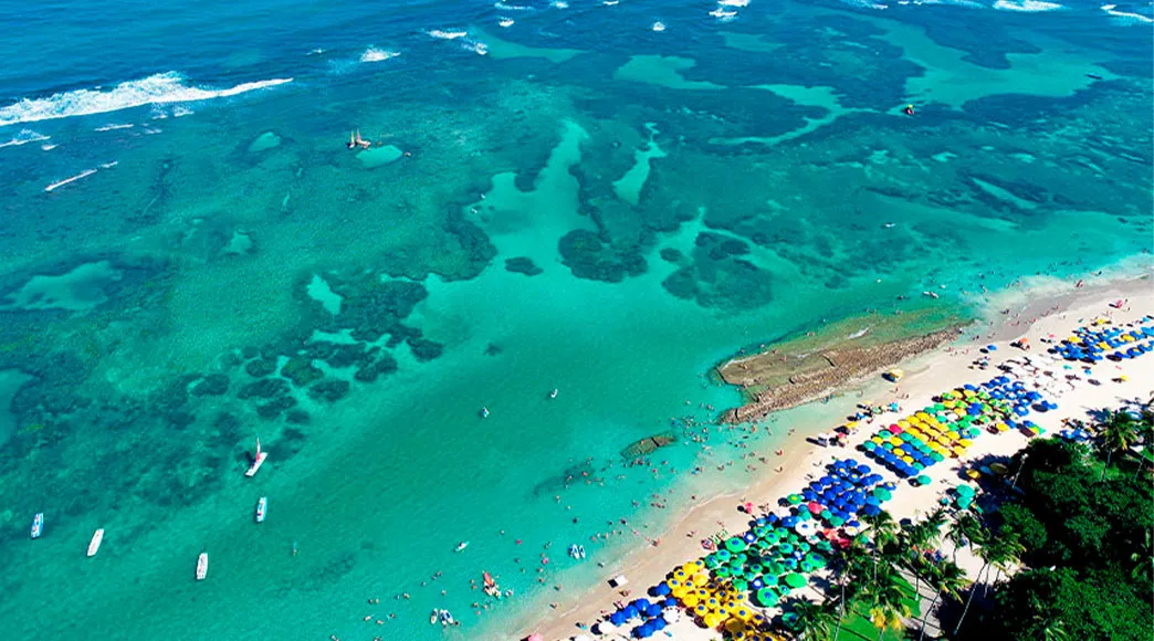 Vista aérea de Porto de Galinhas Vista aérea da praia de Porto de Galinhas, mostrando o mar azul-turquesa com seus recifes e a extensa faixa de areia completamente tomada por centenas de guarda-sóis coloridos e banhistas.