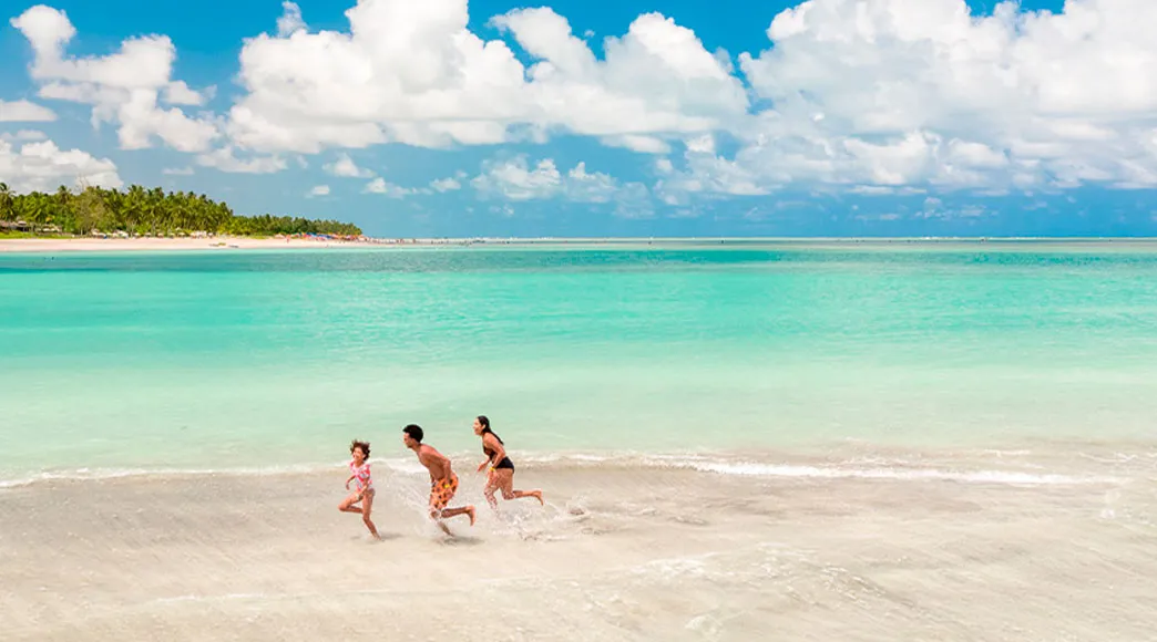 Família brincando na Praia de Antunes Vista aérea de uma família, com pai, mãe e uma filha, correndo pela beira da água na Praia de Antunes. O mar é calmo e de um tom verde-água cristalino, e ao fundo, uma extensa faixa de coqueiros decora a orla sob um céu azul com nuvens.