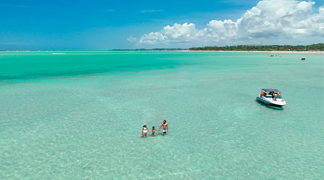 Família nas piscinas naturais de Xaréu Vista aérea de uma família com uma criança em pé no mar raso e cristalino das piscinas naturais da Praia de Xaréu. Uma lancha está próxima, e ao fundo, a orla da praia se estende sob um céu azul com nuvens.