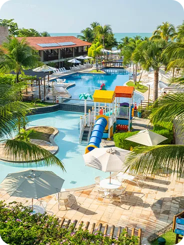 Resort com piscina Vista das piscinas do resort com parque aquático infantil colorido, toboágua azul, espreguiçadeiras, palmeiras e o mar ao fundo.