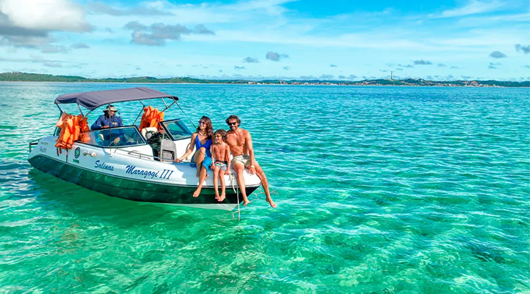 Família em passeio de lancha em Maragogi Uma família com pai, mãe e filho sentados na proa de uma lancha, sorrindo, durante um passeio nas águas cristalinas de cor verde-esmeralda das Galés de Maragogi, com a orla ao fundo.