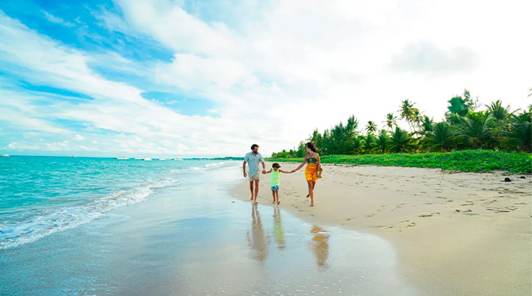 Família caminhando na Praia de Burgalhau Pai, mãe e filho caminham de mãos dadas na beira do mar na Praia de Burgalhau. A areia molhada reflete o céu azul, e a praia é margeada por uma fileira de coqueiros ao fundo.