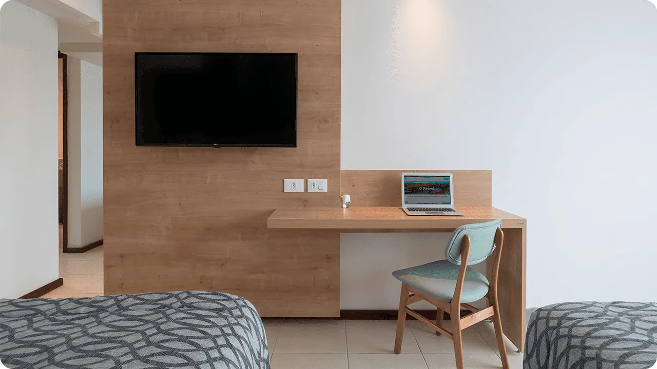 interior da acomodação superior Painel de madeira que fica em frente às camas, com TV a cabo, mesa, cadeira e espelho ao lado.