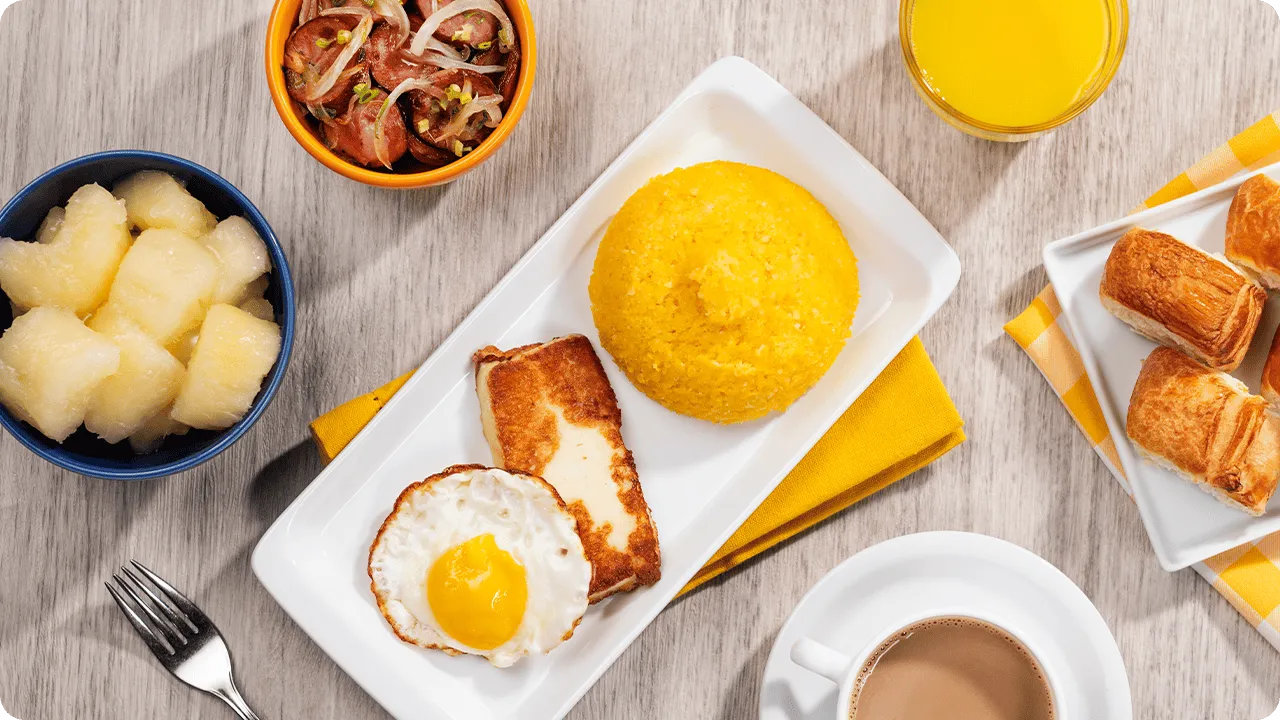 Café da manhã all inclusive Prato com cuscuz amarelo, ovo frito e queijo coalho; ao lado, macaxeira cozida, linguiça acebolada, folhados, suco de laranja e café com leite — café da manhã do Salinas Maragogi all inclusive.