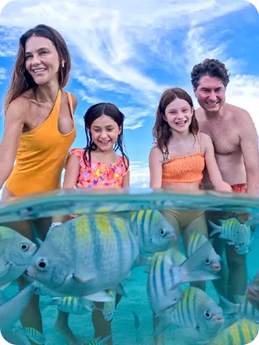 Familia en piscinas naturales Fotografía sumergida y subacuática que muestra a una familia sonriente interactuando con bancos de peces de colores en aguas cristalinas, con los pies de la familia visibles en el fondo marino.