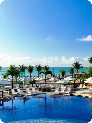 Resort beira-mar com piscina e coqueiros Piscina redonda cercada por coqueiros, guarda-sóis e espreguiçadeiras, com o mar azul e céu aberto ao fundo em um resort à beira-mar.
