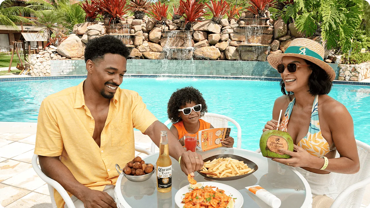 All inclusive à beira da piscina Família sentada à mesa na área da piscina do Salinas Maragogi, com cascata e jardim ao fundo; na mesa, camarões ao molho, batata frita, bolinhos e bebidas como cerveja, suco e água de coco.