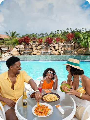 All inclusive à beira da piscina Família sentada à mesa na área da piscina do Salinas Maragogi, com cascata e jardim ao fundo; na mesa, camarões ao molho, batata frita, bolinhos e bebidas como cerveja, suco e água de coco.