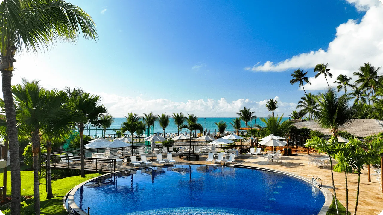 Resort beira-mar com piscina e coqueiros Piscina redonda cercada por coqueiros, guarda-sóis e espreguiçadeiras, com o mar azul e céu aberto ao fundo em um resort à beira-mar.