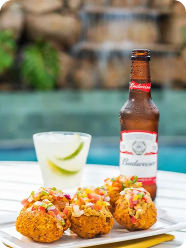 Bebidas e petiscos à beira d'água Prato com bolinhos crocantes cobertos por vinagrete ao lado de uma cerveja Budweiser e um drink cítrico com gelo sobre mesa branca; ao fundo, cascata e piscina do Salinas Maragogi.