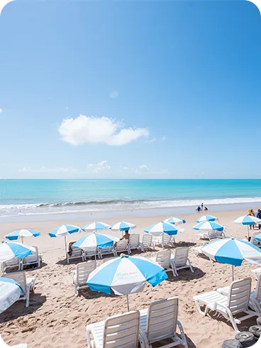 Praia de Ipioca com guarda-sóis e mar turquesa Cadeiras e guarda-sóis azuis e brancos na areia da Praia de Ipioca, com mar turquesa e céu azul ao fundo.