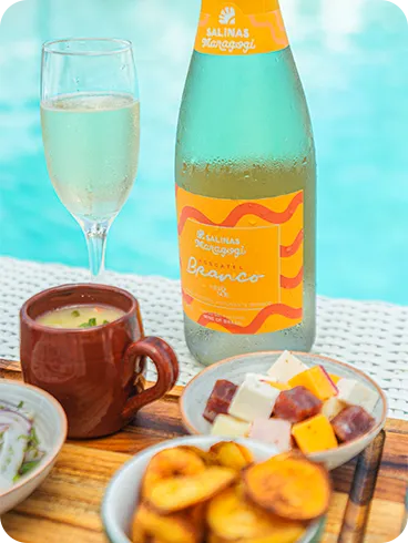 Vino espumoso Salinas Maragogi y aperitivos junto a la piscina Una botella de vino blanco espumoso Moscatel Salinas Maragogi junto a una copa y aperitivos, incluyendo una porción de camarones, queso en cubos y ceviche, con la piscina de fondo.