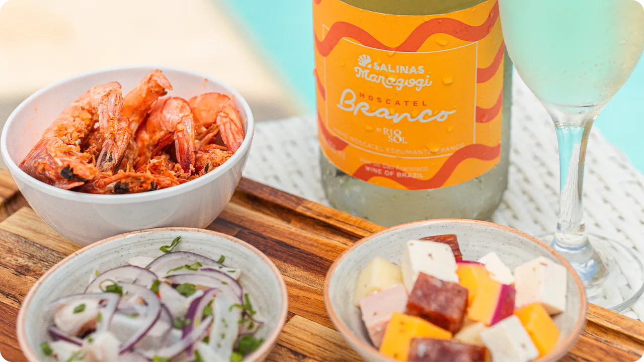 Vino espumoso Salinas Maragogi y aperitivos junto a la piscina Una botella de vino blanco espumoso Moscatel Salinas Maragogi junto a una copa y aperitivos, incluyendo una porción de camarones, queso en cubos y ceviche, con la piscina de fondo.