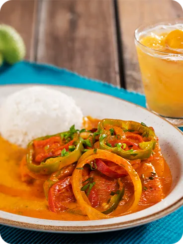 All inclusive com sabor brasileiro Prato de moqueca com arroz branco, molho cremoso alaranjado e rodelas de pimentão e tomate, acompanhado de caipirinha em copo baixo sobre toalha azul.