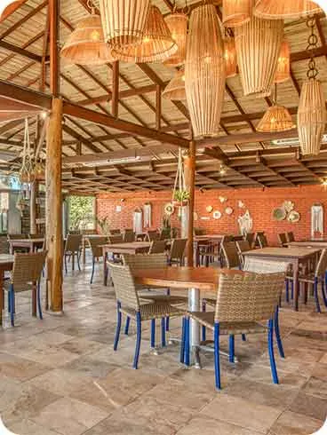Restaurante no Salinas Maceió Interior de um dos restaurantes do Salinas Maceió com mesas de madeira, cadeiras de fibra, luminárias de palha e vista para o mar ao fundo.