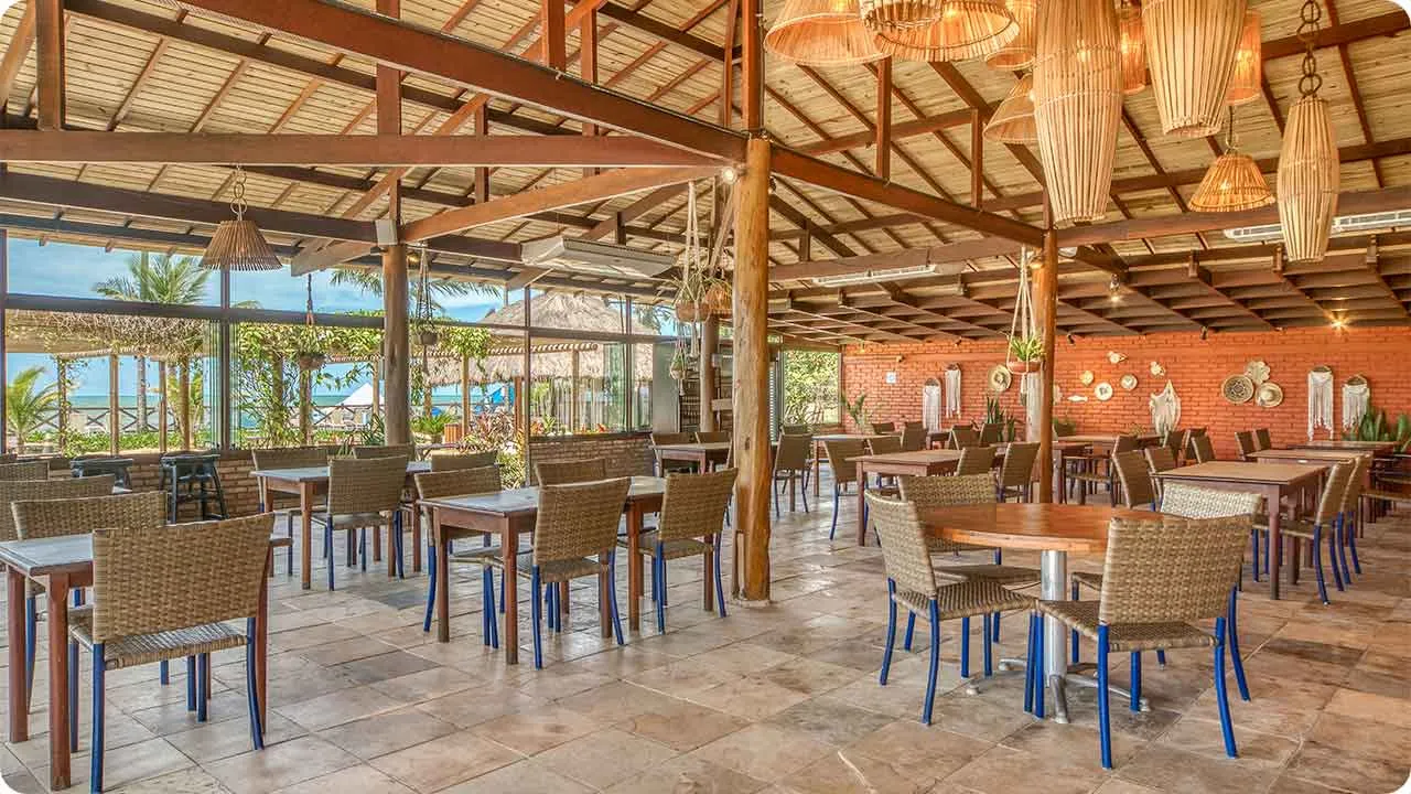 Restaurante no Salinas Maceió Interior de um dos restaurantes do Salinas Maceió com mesas de madeira, cadeiras de fibra, luminárias de palha e vista para o mar ao fundo.
