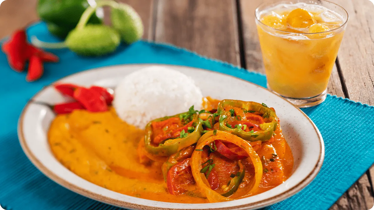 All inclusive com sabor brasileiro Prato de moqueca com arroz branco, molho cremoso alaranjado e rodelas de pimentão e tomate, acompanhado de caipirinha em copo baixo sobre toalha azul.