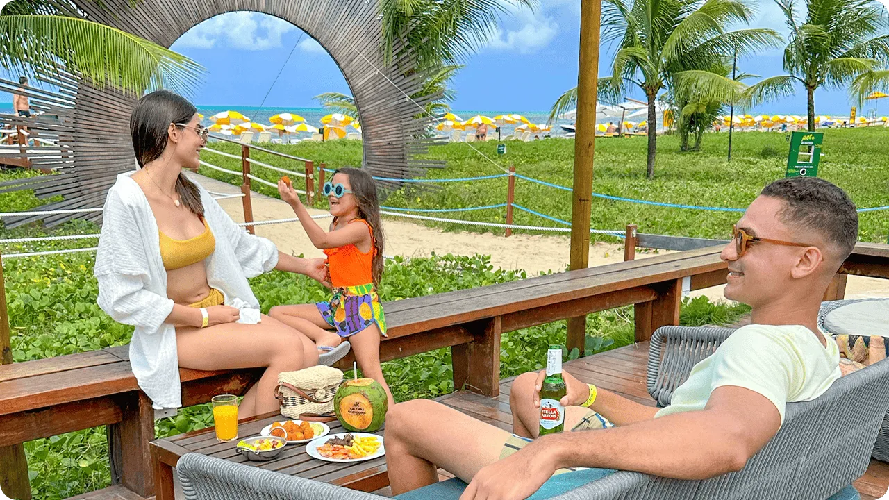 All inclusive à beira-mar Família aproveitando o espaço externo do resort, sentada em sofás de madeira com vista para o mar e coqueiros, mesa com petiscos, coco e bebidas.
