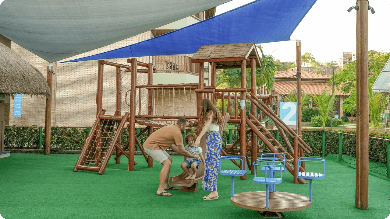 Lazer infantil no Salinas Maceió Família brincando no parquinho do resort, com escorregador e estrutura de madeira sobre piso macio e cobertura de sombreamento.