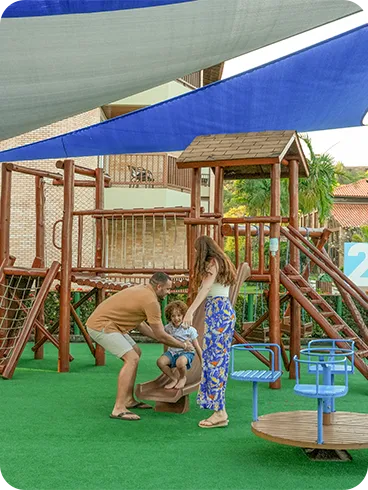 Lazer infantil no Salinas Maceió Família brincando no parquinho do resort, com escorregador e estrutura de madeira sobre piso macio e cobertura de sombreamento.