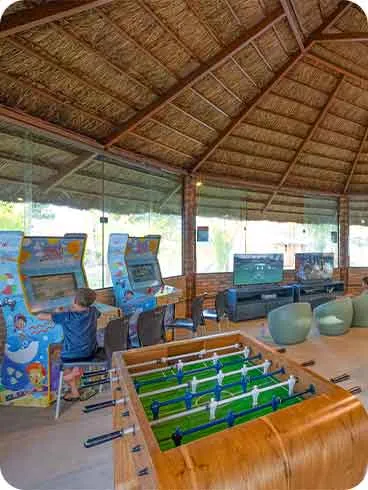 Sala de jogos no Salinas Maceió Espaço coberto com telhado de palha e paredes de vidro, com mesas de pingue-pongue e pebolim, arcades, videogames e air hockey, crianças jogando e se divertindo.