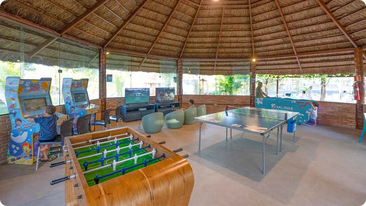 Sala de jogos no Salinas Maceió Espaço coberto com telhado de palha e paredes de vidro, com mesas de pingue-pongue e pebolim, arcades, videogames e air hockey, crianças jogando e se divertindo.