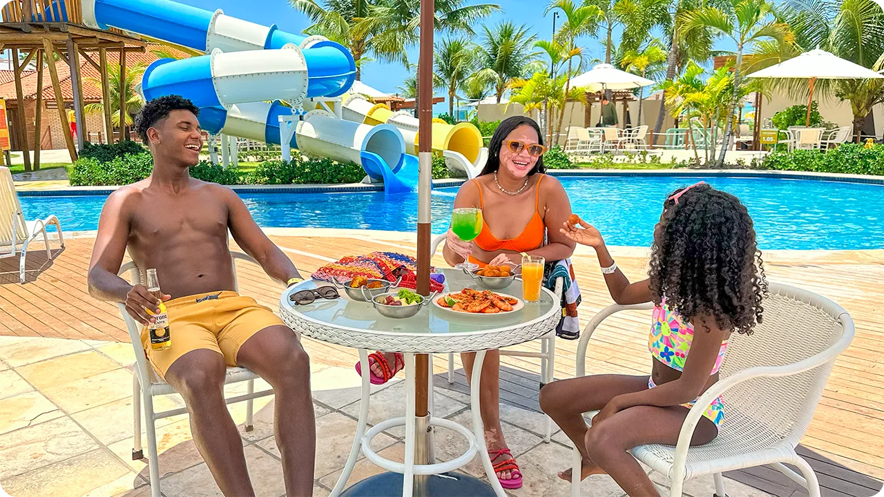 Familia en la zona de la piscina de Salinas Maceió Padre, madre e hija disfrutan de un refrigerio en una mesa junto a la piscina del resort, con aperitivos y bebidas. Al fondo, el parque acuático con sus grandes toboganes azules y blancos completa la escena de vacaciones familiares.