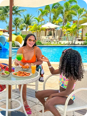 Familia en la zona de la piscina de Salinas Maceió Padre, madre e hija disfrutan de un refrigerio en una mesa junto a la piscina del resort, con aperitivos y bebidas. Al fondo, el parque acuático con sus grandes toboganes azules y blancos completa la escena de vacaciones familiares.