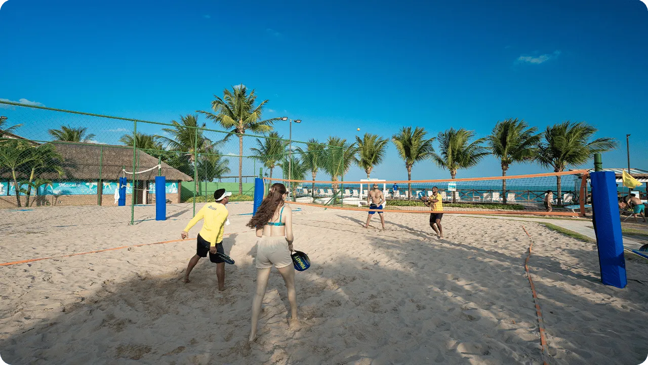 Resort com diversão Grupo jogando beach tênis em quadra de areia no resort, com palmeiras ao fundo, céu azul e clima praiano.