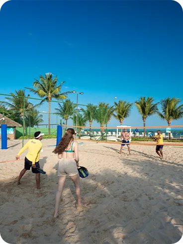 Resort com diversão Grupo jogando beach tênis em quadra de areia no resort, com palmeiras ao fundo, céu azul e clima praiano.