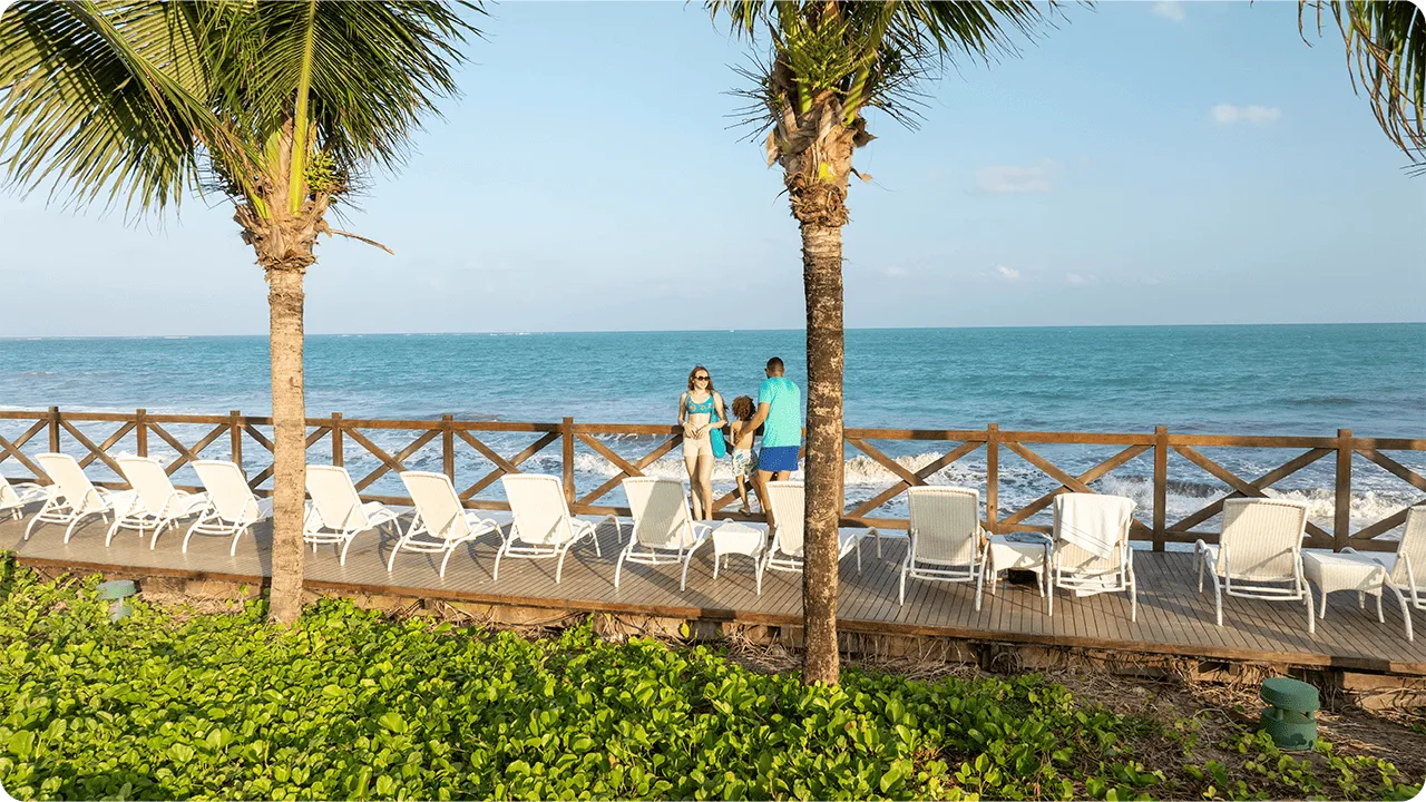 Resort beira-mar Deck de madeira à beira-mar com espreguiçadeiras brancas alinhadas entre palmeiras; ao centro, família com uma criança observa o mar azul sob céu claro.