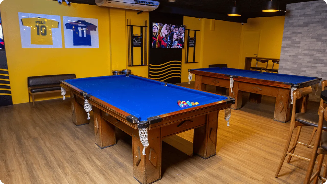 Bar no salinas maceió Salão do bar com duas mesas de sinuca de pano azul, paredes amarelas, camisas de futebol emolduradas, TV transmitindo jogo, bancos altos e piso de madeira.