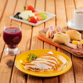 Café da manhã no Salinas Maceió Mesa de café da manhã no resort, com tapioca doce, pães, frios, frutas frescas, suco e café, servidos em uma mesa de madeira.